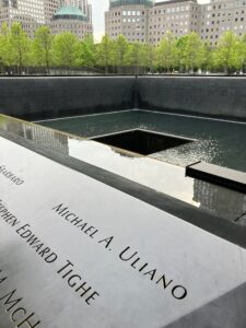 Photo of the 9/11 Memorial, with people's names etched in metal in the foreground, and the pool with the "footprint" of one of the towers behind it, and the buildings of the city behind that.