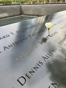 Photo of the 9/11 Memorial, with names etched in metal with a single flower left for someone, with the waterfalls and pools of the Memorial in the background.